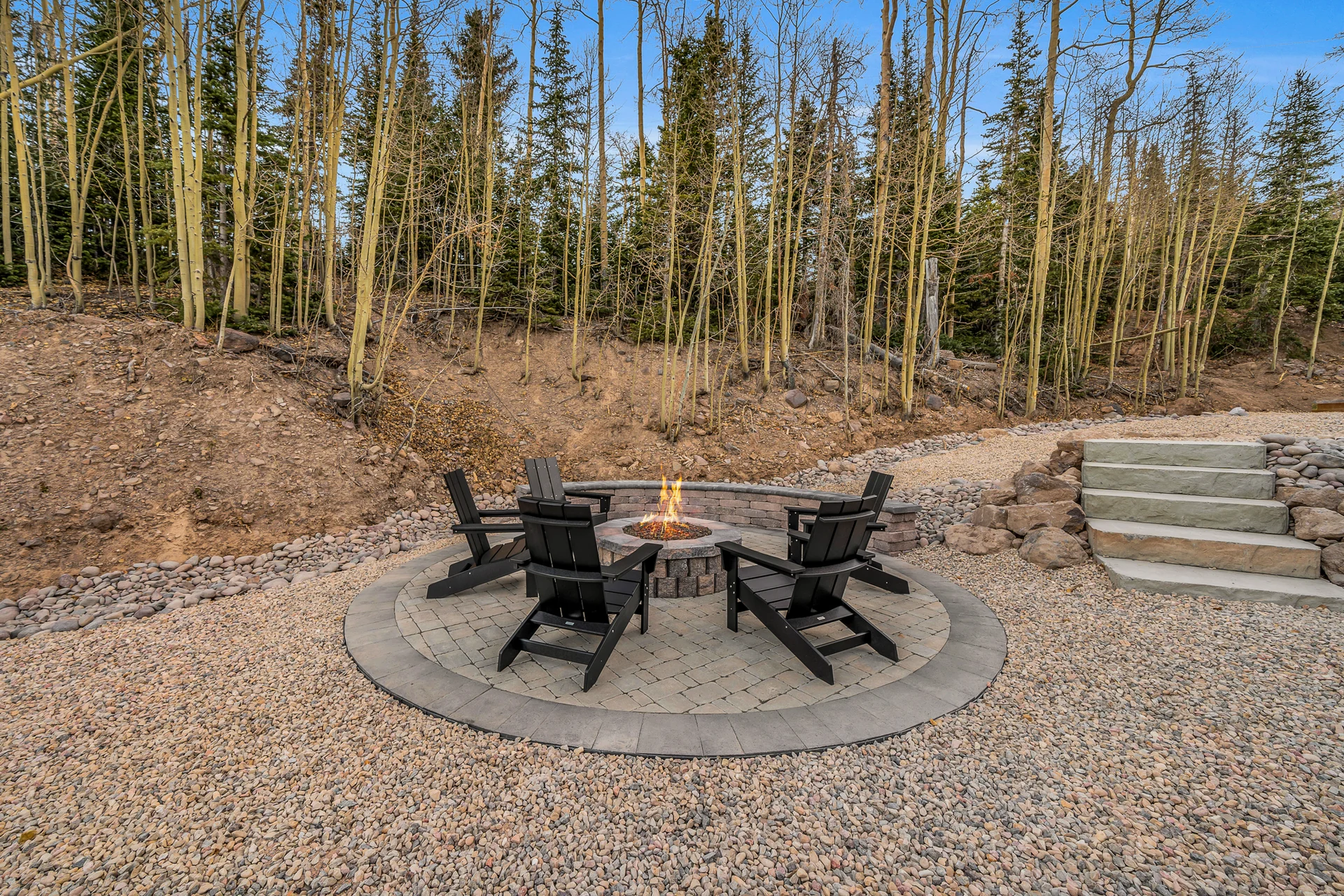 Black Adirondack chairs surround a circular stone fire pit with a visible flame. Stone steps, gravel and trees sit behind the paved seating area.
