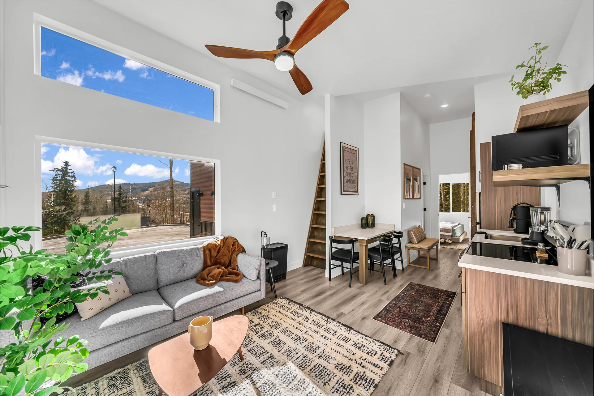 Sofa and coffee table beside tall windows, a compact dining table, kitchen, steep wooden loft access and a doorway into the queen bedroom.