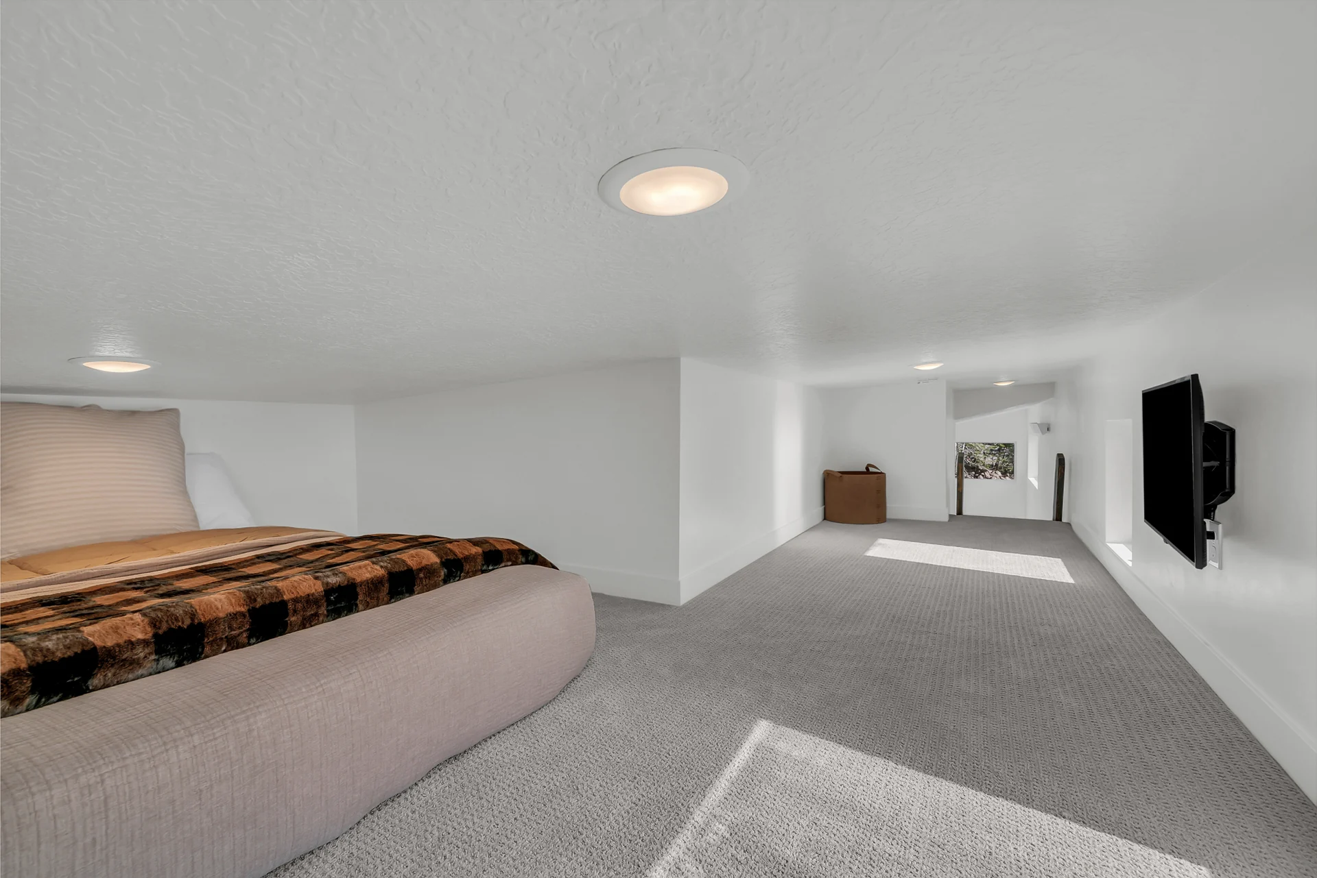 Reverse view of a low-ceiling loft with part of a bed at left, carpeted floor, a black TV at right and the loft opening at the back.